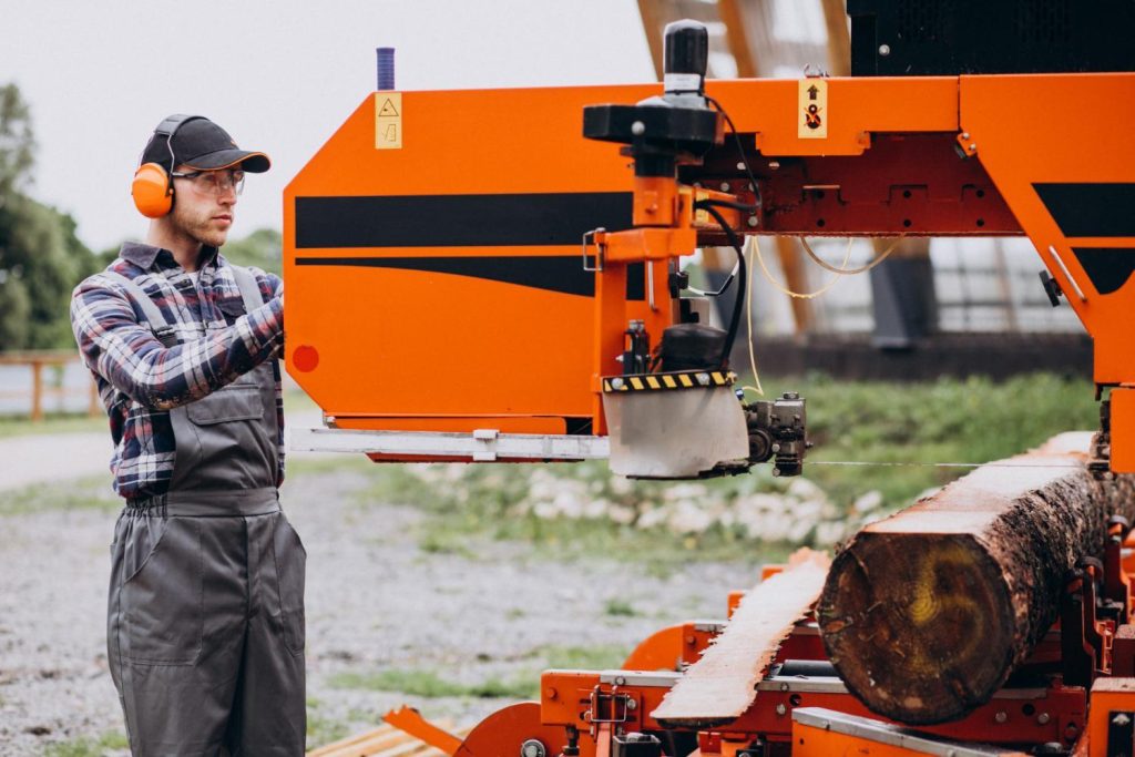 Worker wearing protective gear operating a portable sawmill to cut large logs into manageable firewood sections outdoors