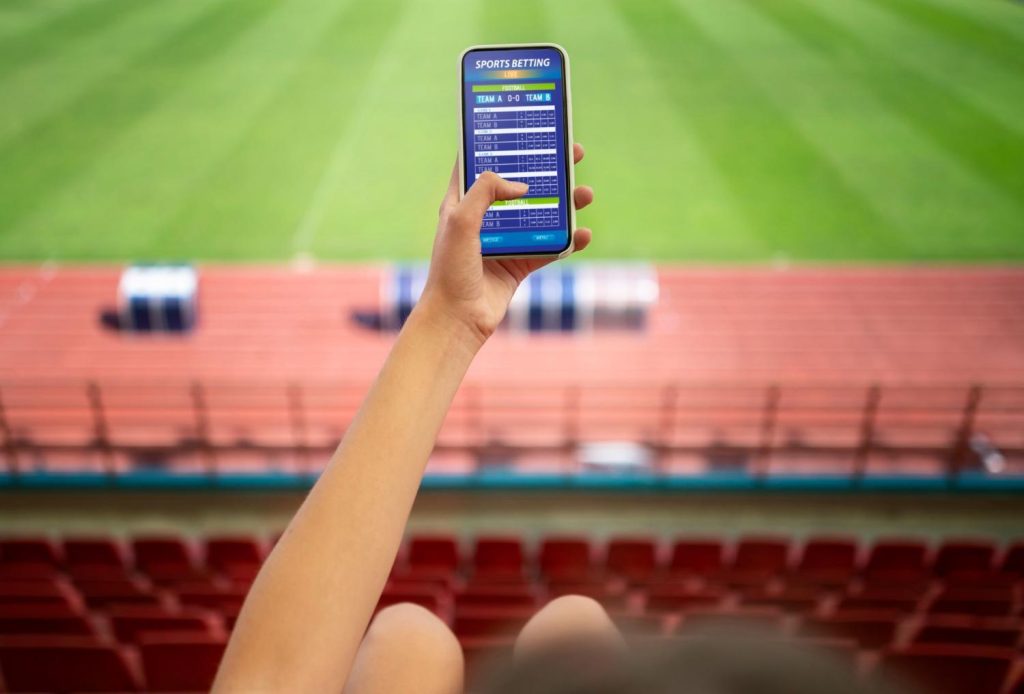 Person holding a smartphone displaying a live sports betting app while seated in a stadium, showing modern mobile betting in action