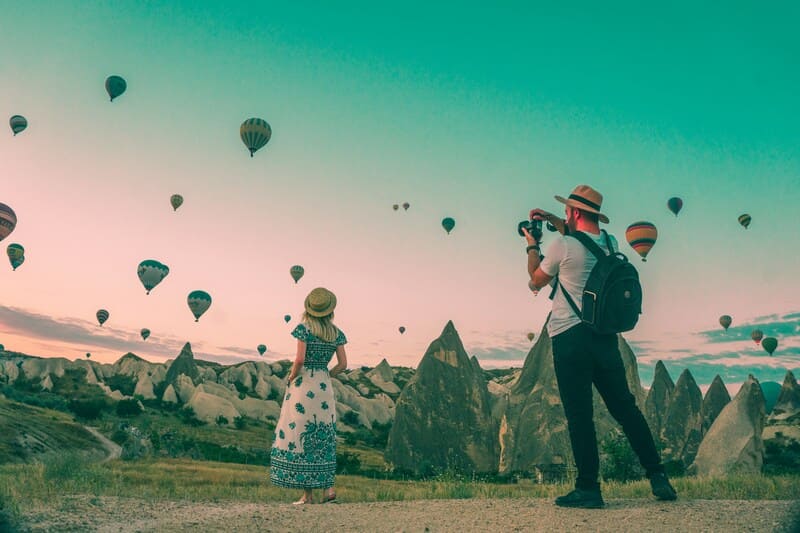Two travelers with camera and backpack watching hot air balloons over rocky landscape at sunrise